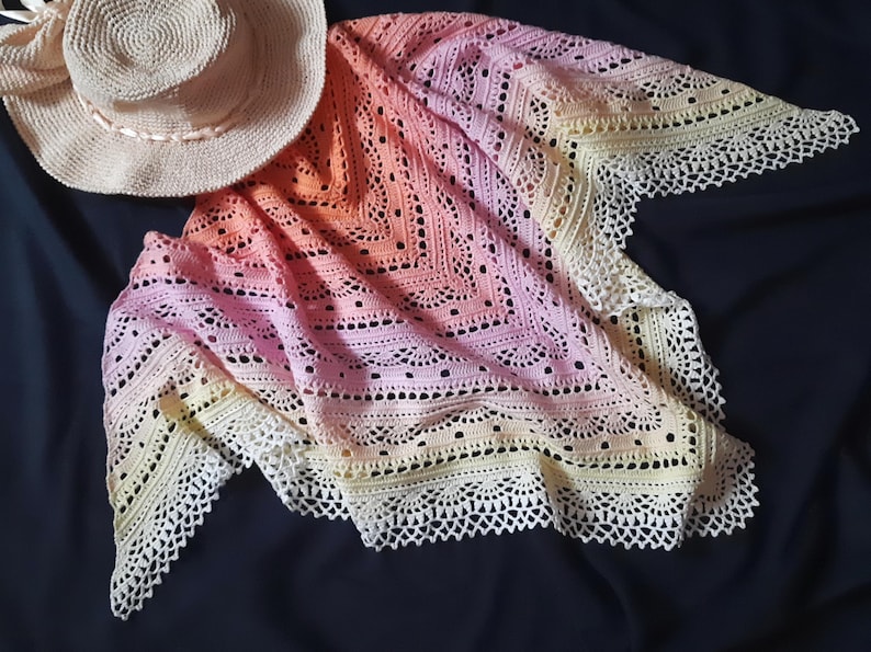 May include: A handmade, crochet shawl in a gradient of colors, from pale yellow to peach and pink, with a delicate, lacy border. A straw hat with a ribbon sits beside the shawl. The shawl is a beautiful accessory.