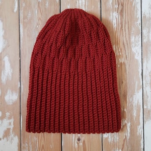 May include: A red knitted beanie hat with a ribbed texture. The hat is photographed against a light wood background.