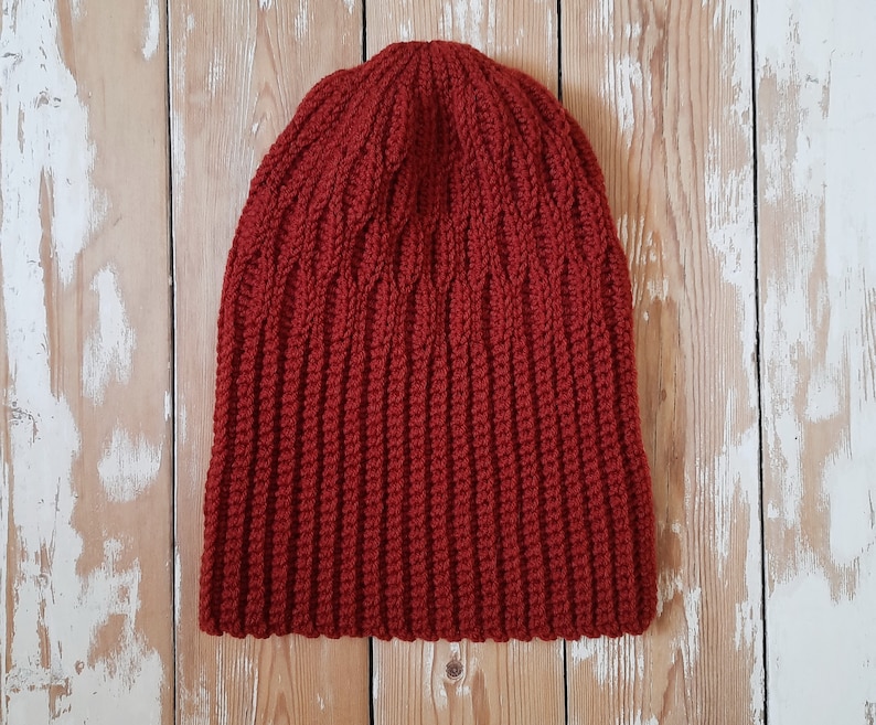 May include: A red knitted beanie hat with a ribbed texture. The hat is photographed against a light wood background.