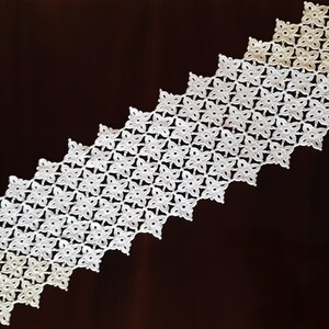 May include: White crocheted table runner with a graduated pattern of small flowers. The runner is on a dark brown wooden surface.