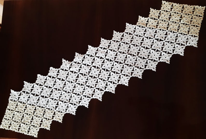 May include: White crocheted table runner with a graduated pattern of small flowers. The runner is on a dark brown wooden surface.