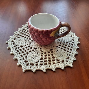 May include: A white ceramic cup with a grape design sits on a white crocheted doily.