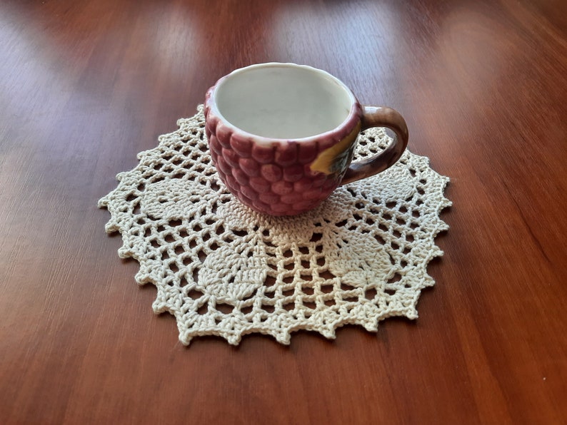 May include: A white ceramic cup with a grape design sits on a white crocheted doily.