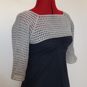 K&ouml;nnte beinhalten: Ein hellgrauer, offen gewebter H&auml;kel-Bolero mit Dreiviertel&auml;rmeln. Der Bolero wird auf einer roten Schneiderpuppe &uuml;ber einem dunkelblauen Kleid pr&auml;sentiert. Das H&auml;kelmuster erzeugt eine zarte, luftige Textur. Der Ausschnitt ist quadratisch.