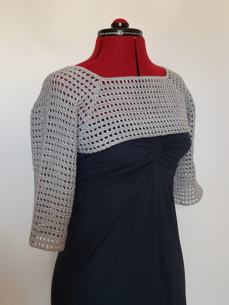 K&ouml;nnte beinhalten: Ein hellgrauer, offen gewebter H&auml;kel-Bolero mit Dreiviertel&auml;rmeln. Der Bolero wird auf einer roten Schneiderpuppe &uuml;ber einem dunkelblauen Kleid pr&auml;sentiert. Das H&auml;kelmuster erzeugt eine zarte, luftige Textur. Der Ausschnitt ist quadratisch.