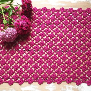 Puede incluir: Un mantel de ganchillo rosa con un delicado patr&oacute;n floral. El mantel est&aacute; hecho con un hilo fino y tiene un borde festoneado.