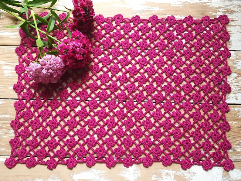 Puede incluir: Un mantel de ganchillo rosa con un delicado patr&oacute;n floral. El mantel est&aacute; hecho con un hilo fino y tiene un borde festoneado.