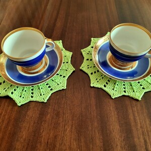 May include: Two blue and gold teacups with white interiors, sitting on green crocheted doilies.