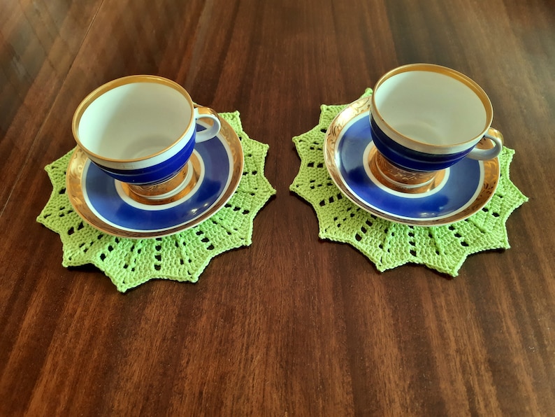 May include: Two blue and gold teacups with white interiors, sitting on green crocheted doilies.