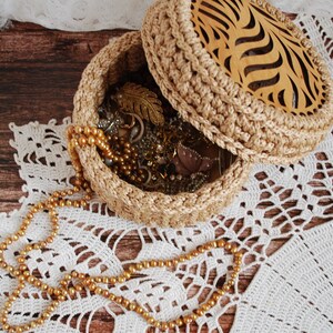 Pu&ograve; includere: Una scatola per gioielli rotonda in crochet con un coperchio in legno con un motivo ondulato intagliato. La scatola &egrave; piena di gioielli d'oro e perle e si trova su un pizzo all'uncinetto bianco.