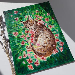 Puede incluir: Una pintura de un gato atigrado marr&oacute;n durmiendo en un campo de flores rosas. El gato est&aacute; acurrucado de lado con los ojos cerrados. Las flores est&aacute;n esparcidas alrededor del gato, y el fondo es un exuberante campo verde.