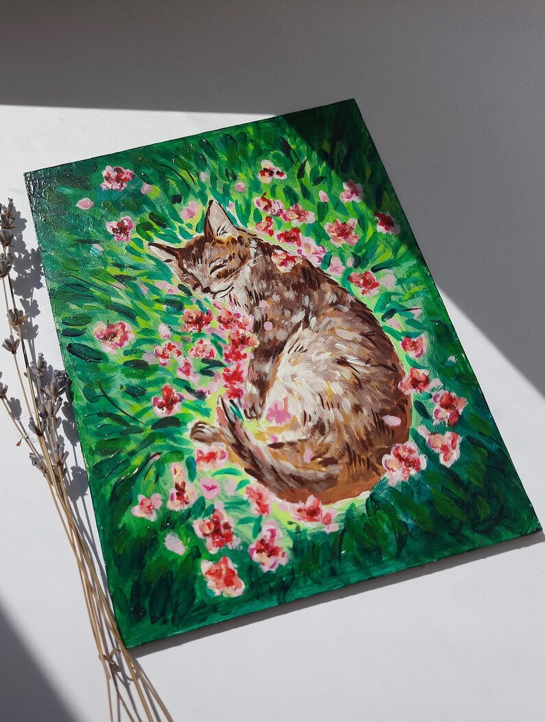 Puede incluir: Una pintura de un gato atigrado marr&oacute;n durmiendo en un campo de flores rosas. El gato est&aacute; acurrucado de lado con los ojos cerrados. Las flores est&aacute;n esparcidas alrededor del gato, y el fondo es un exuberante campo verde.