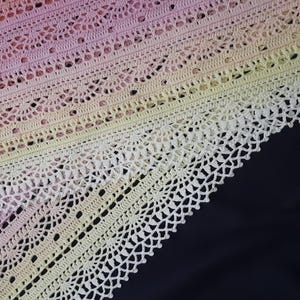 May include: Close-up of a crocheted shawl with a gradient of colors, transitioning from pink to yellow. The intricate lace pattern features rows of delicate openwork and scalloped edges, set against a dark background.