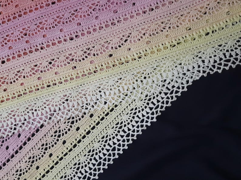 May include: Close-up of a crocheted shawl with a gradient of colors, transitioning from pink to yellow. The intricate lace pattern features rows of delicate openwork and scalloped edges, set against a dark background.