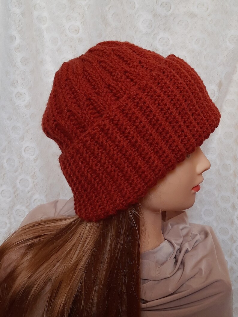 May include: A rust-colored knitted beanie with a fold-over brim. The beanie is made with a thick yarn and has a ribbed texture.