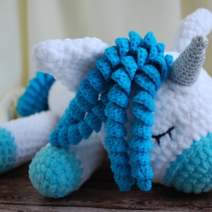May include: A white crocheted unicorn with a blue yarn mane and hooves. The unicorn has a silver horn and closed eyes.