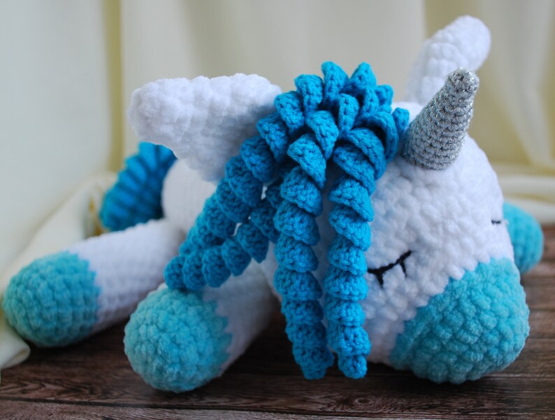 May include: A white crocheted unicorn with a blue yarn mane and hooves. The unicorn has a silver horn and closed eyes.