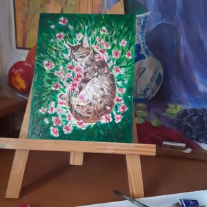 Puede incluir: Una pintura en un caballete de madera representa un gato marr&oacute;n y blanco durmiendo en una cama de flores rosas sobre un fondo verde.