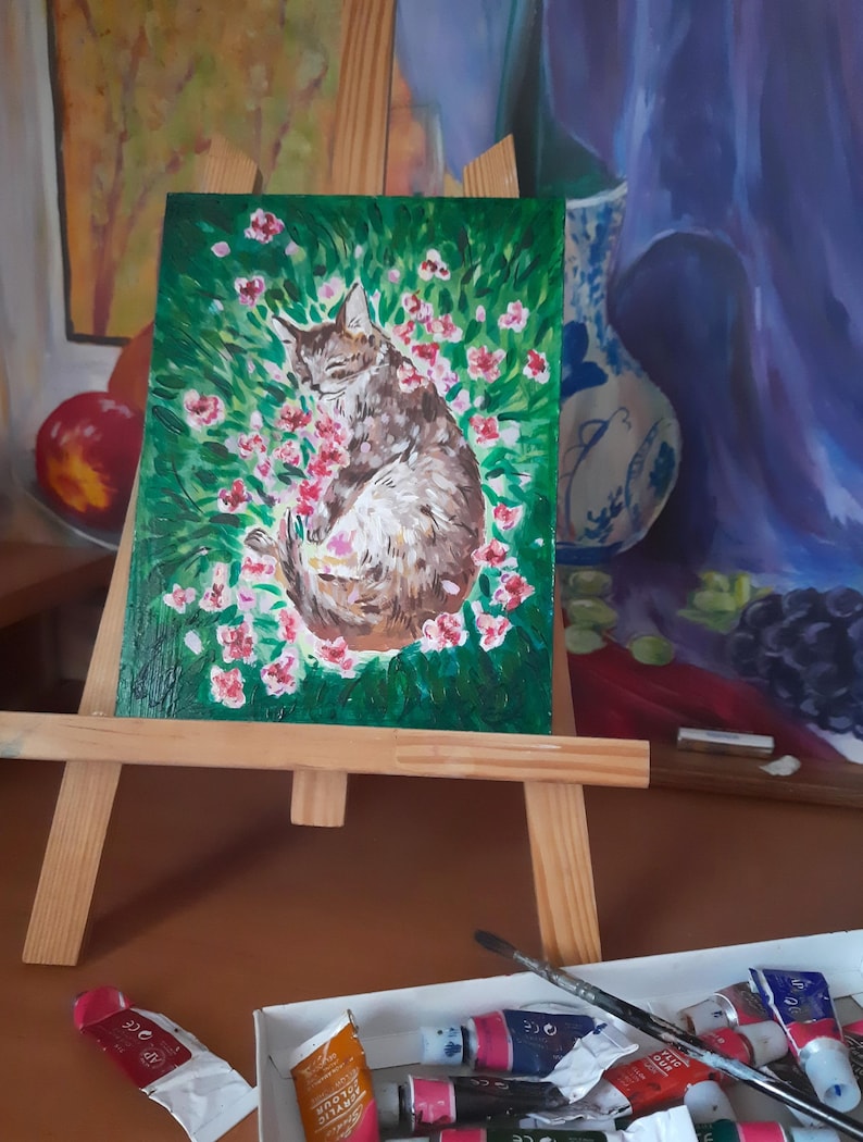 Puede incluir: Una pintura en un caballete de madera representa un gato marr&oacute;n y blanco durmiendo en una cama de flores rosas sobre un fondo verde.