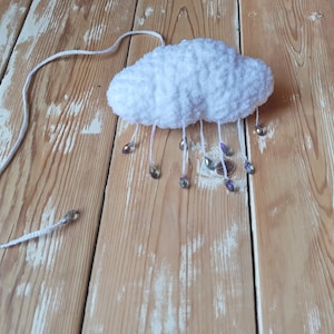 Puede incluir: Una nube blanca y esponjosa de ganchillo con cuentas de cristal colgantes que simulan gotas de lluvia. La nube est&aacute; suspendida por un cord&oacute;n blanco. El objeto artesanal se encuentra sobre un fondo de madera clara.