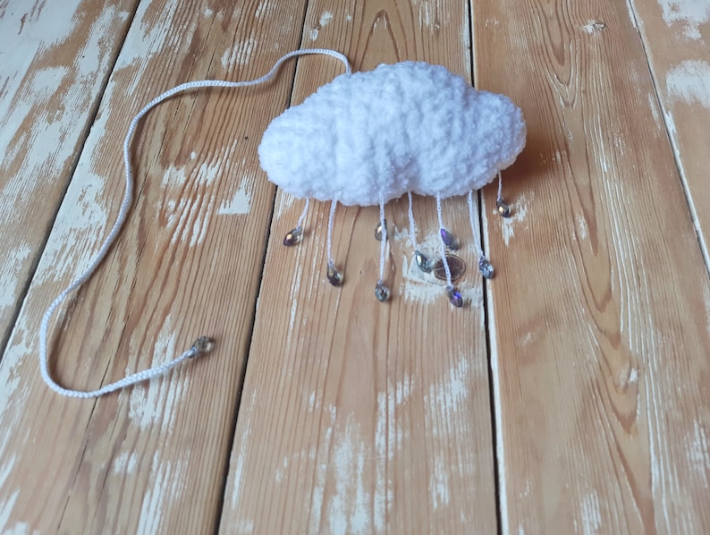 Puede incluir: Una nube blanca y esponjosa de ganchillo con cuentas de cristal colgantes que simulan gotas de lluvia. La nube est&aacute; suspendida por un cord&oacute;n blanco. El objeto artesanal se encuentra sobre un fondo de madera clara.