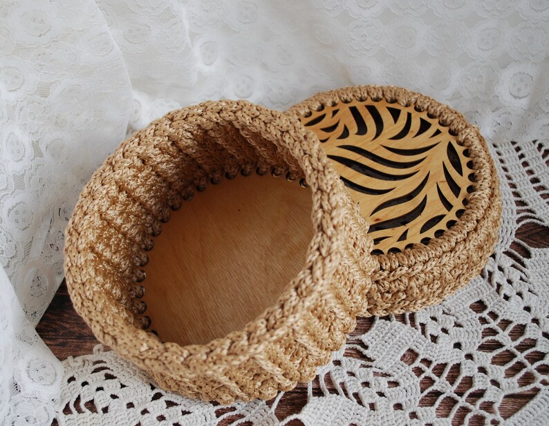 Pu&ograve; includere: Una scatola di immagazzinaggio rotonda in crochet con un coperchio in legno con un design a foglia ritagliata. La scatola &egrave; beige e il coperchio &egrave; in legno naturale.