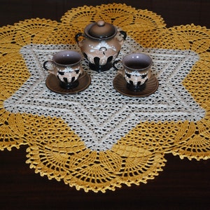 May include: A white and yellow crocheted doily with a star-shaped centre. Three small cups and a teapot with floral designs are placed on the doily.
