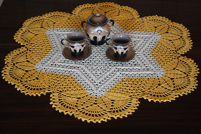 May include: A white and yellow crocheted doily with a star-shaped centre. Three small cups and a teapot with floral designs are placed on the doily.