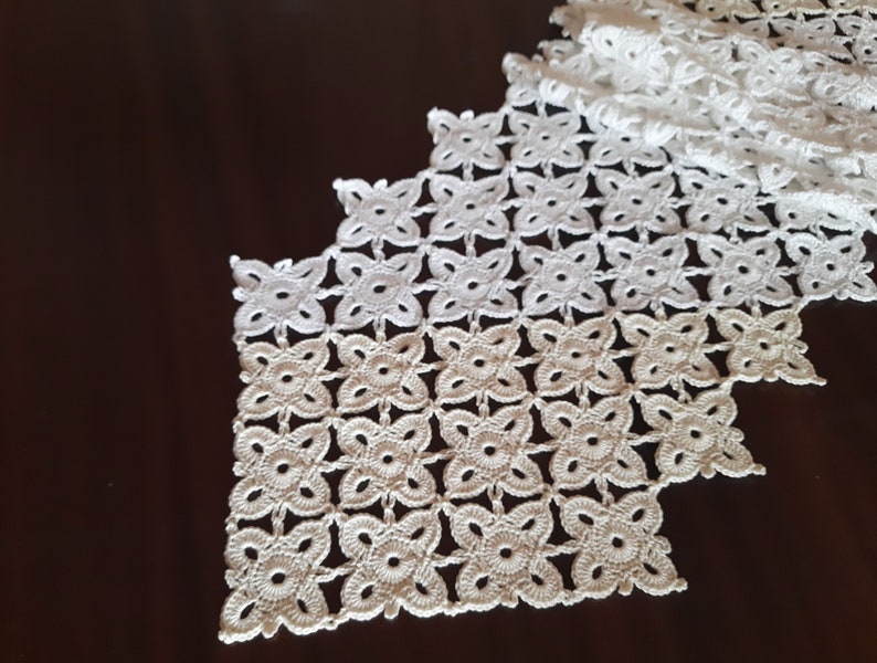 May include: White crocheted table runner with a delicate floral pattern. The runner is made of a fine thread and has a scalloped edge.