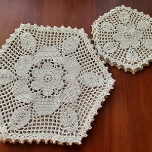 May include: Two crocheted doilies in a beige color. The larger doily is hexagonal in shape and the smaller doily is round. Both doilies have a delicate floral pattern.