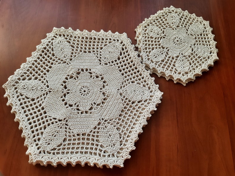 May include: Two crocheted doilies in a beige color. The larger doily is hexagonal in shape and the smaller doily is round. Both doilies have a delicate floral pattern.