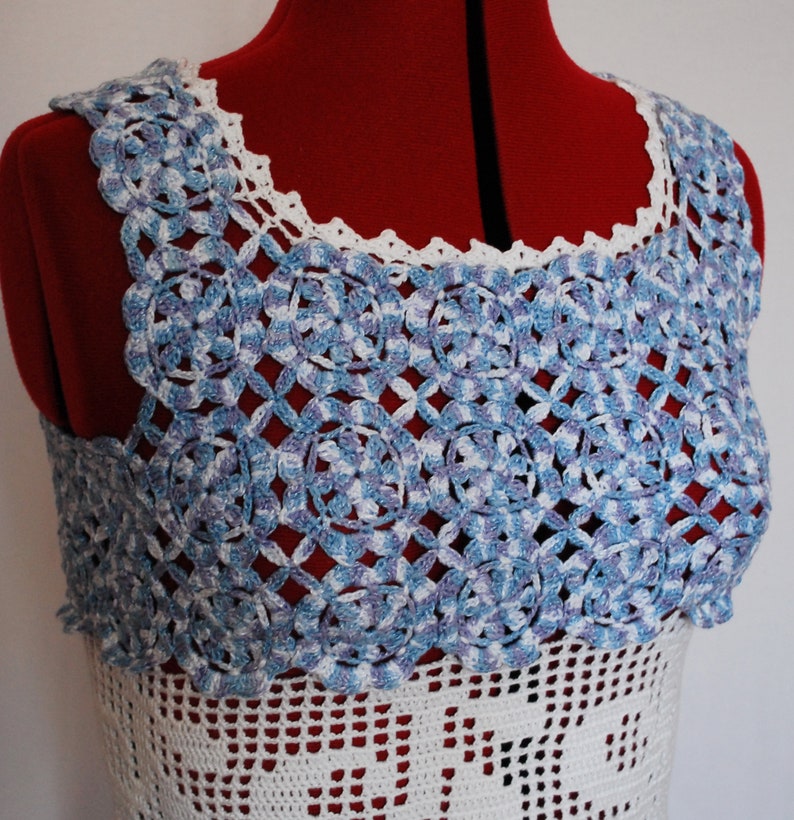 Puede incluir: Un top de crochet blanco con un borde de encaje de crochet azul y blanco alrededor del escote. El top es sin mangas y tiene un ajuste holgado.