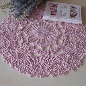 May include: A handmade, light purple crochet doily with a detailed floral design. A book with floral illustrations and Russian text rests on the doily, along with a bundle of dried lavender. The doily is circular and features a central sunburst pattern.