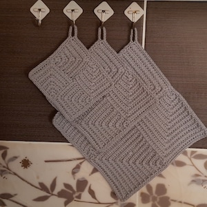 May include: Two gray crocheted pot holders hanging on hooks. The pot holders have a square shape and a textured pattern.