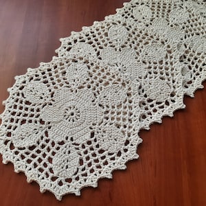 May include: A crocheted table runner with a delicate floral pattern in a cream color. The runner is made of a fine yarn and has a scalloped edge.