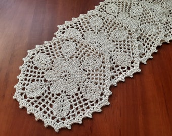 May include: A crocheted table runner with a delicate floral pattern in a cream color. The runner is made of a fine yarn and has a scalloped edge.