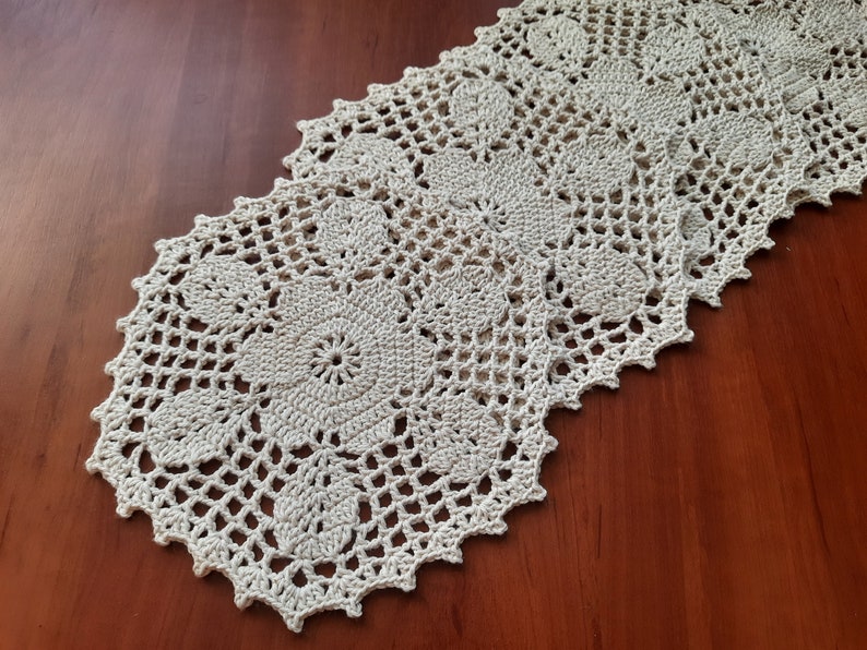 May include: A crocheted table runner with a delicate floral pattern in a cream color. The runner is made of a fine yarn and has a scalloped edge.