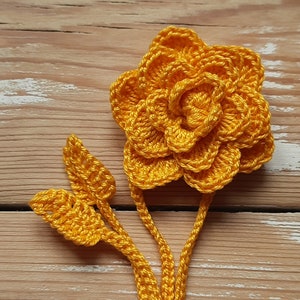 May include: A crocheted yellow flower with two leaves attached to a stem. The flower is made of multiple layers of crocheted petals.