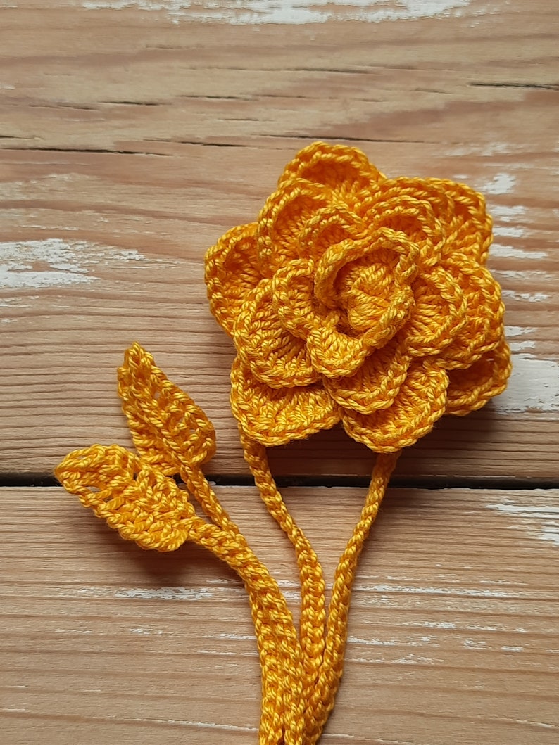 May include: A crocheted yellow flower with two leaves attached to a stem. The flower is made of multiple layers of crocheted petals.
