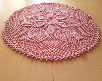 Puede incluir: Un tapete redondo de ganchillo rosa. El diseño presenta un motivo floral central con patrones de pétalos detallados y un borde calado. El color es un rosa suave y apagado, y la textura es intrincada y detallada. Hecho a mano.