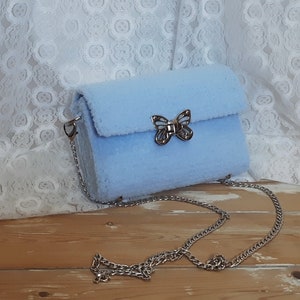 Puede incluir: Un bolso de hombro de color azul claro y peludo con un cierre de mariposa plateado y una correa de cadena plateada.