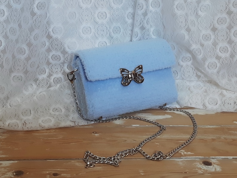 Puede incluir: Un bolso de hombro de color azul claro y peludo con un cierre de mariposa plateado y una correa de cadena plateada.