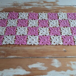 May include: A crocheted table runner with a pink and white checkered pattern. The runner is made of delicate, intricate stitches and has a soft, feminine feel.