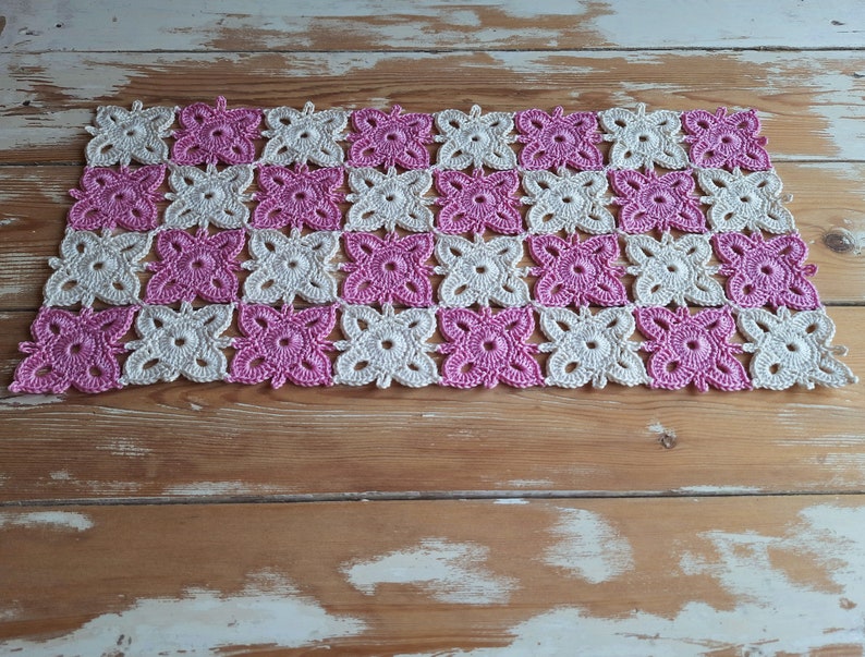 May include: A crocheted table runner with a pink and white checkered pattern. The runner is made of delicate, intricate stitches and has a soft, feminine feel.