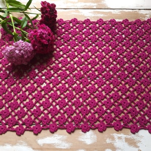 Puede incluir: Un camino de mesa de ganchillo rosa con un patr&oacute;n de diamante. El camino de mesa est&aacute; hecho con un hilo fino y tiene un aspecto delicado y de encaje.