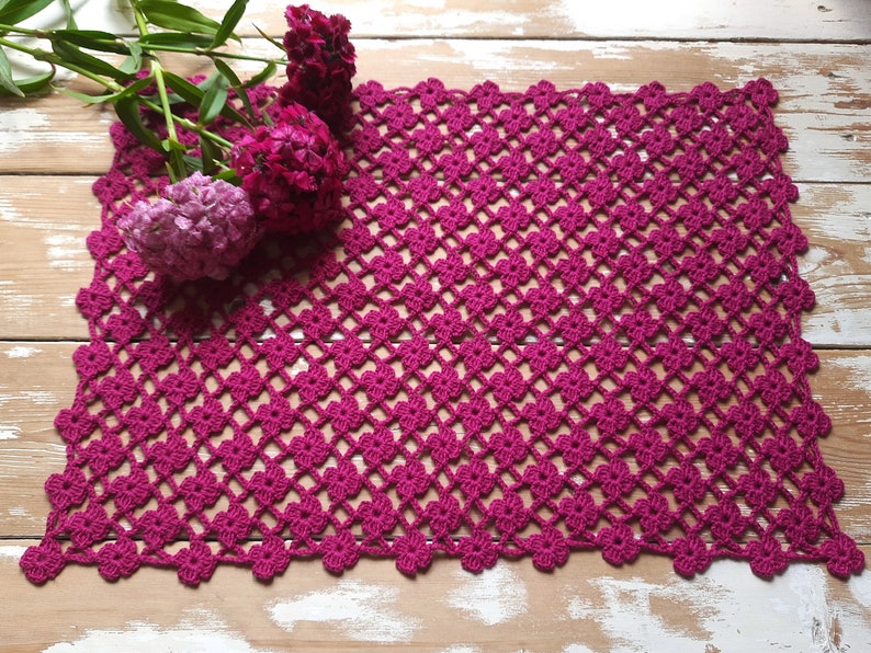 Puede incluir: Un camino de mesa de ganchillo rosa con un patr&oacute;n de diamante. El camino de mesa est&aacute; hecho con un hilo fino y tiene un aspecto delicado y de encaje.
