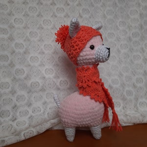 May include: A pink crocheted llama wearing a red crocheted hat and scarf. The llama has white horns and a white tail.