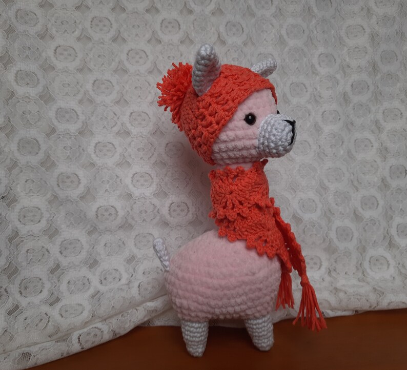 May include: A pink crocheted llama wearing a red crocheted hat and scarf. The llama has white horns and a white tail.