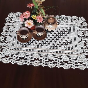 K&ouml;nnte beinhalten: Ein wei&szlig;er geh&auml;kelter Tischl&auml;ufer mit einem zarten Blumendesign. Der L&auml;ufer liegt auf einer dunklen Holzoberfl&auml;che mit zwei Teetassen und einer Teekanne darauf.