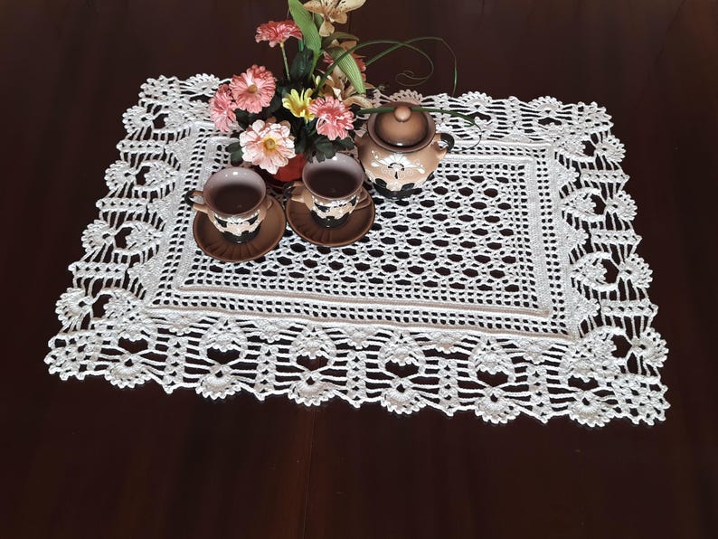 K&ouml;nnte beinhalten: Ein wei&szlig;er geh&auml;kelter Tischl&auml;ufer mit einem zarten Blumendesign. Der L&auml;ufer liegt auf einer dunklen Holzoberfl&auml;che mit zwei Teetassen und einer Teekanne darauf.